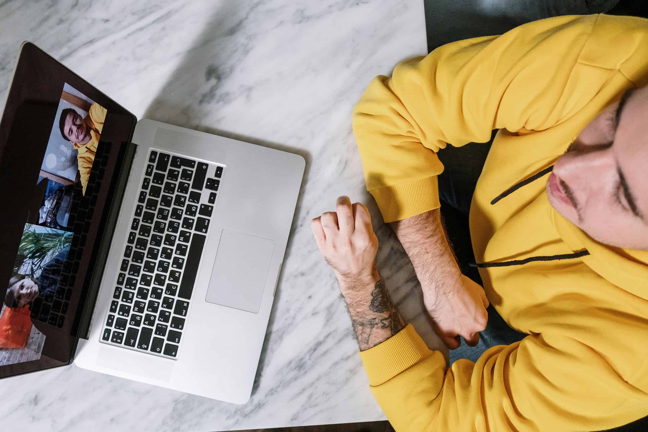 Man in yellow hoodie participating in a virtual meeting using a laptop on a marble table.